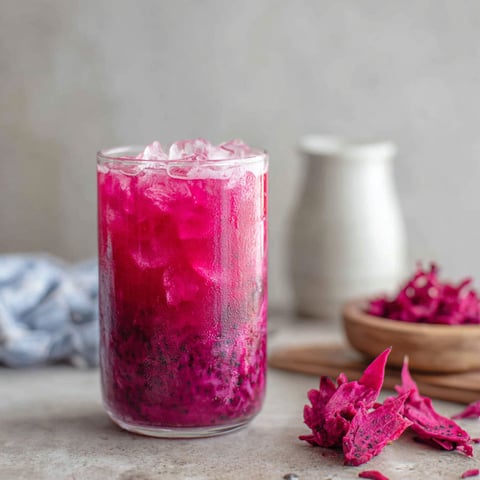 A glass of pink drink with ice and a bowl of fruit next to it.